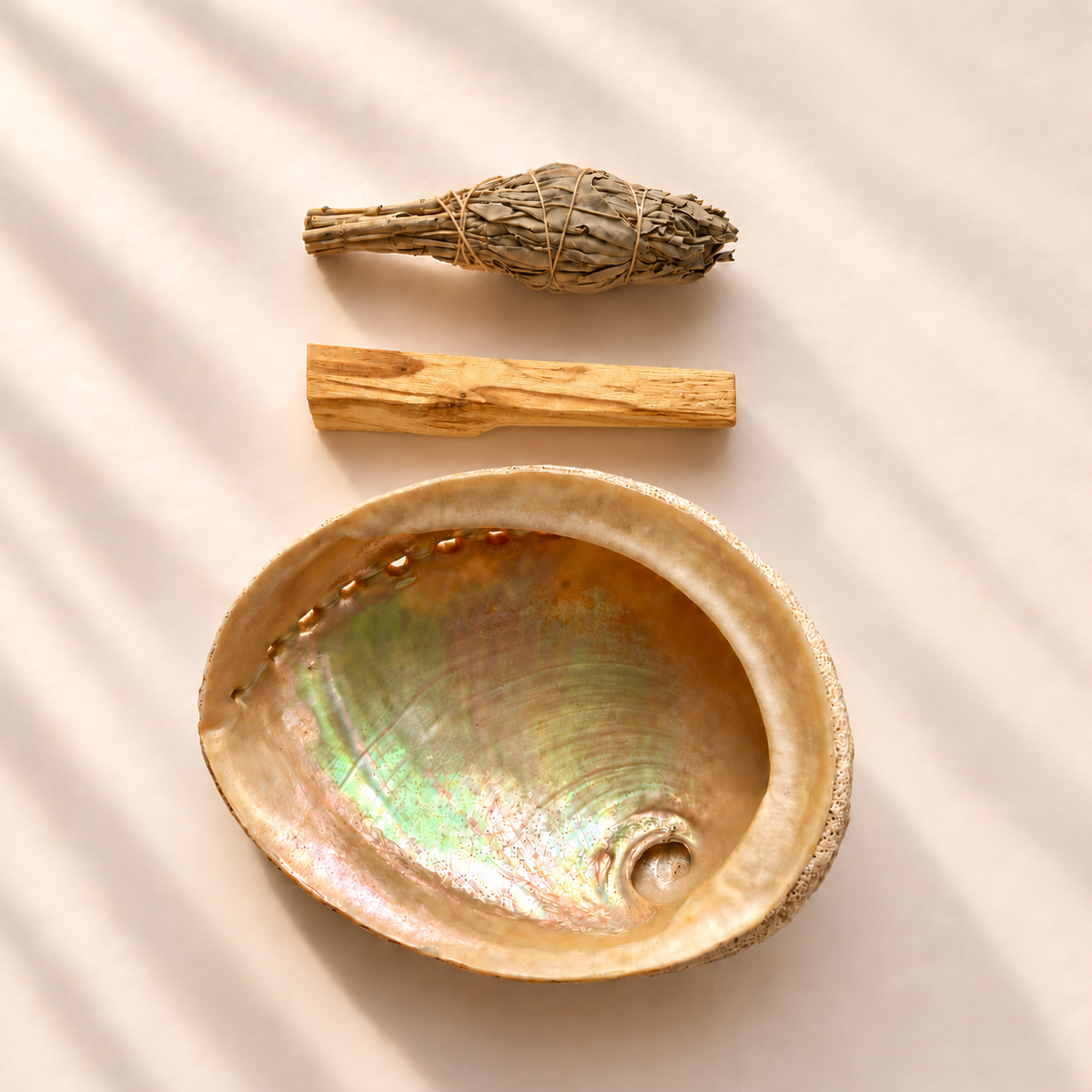 Natural abalone shell with an iridescent interior, dried sage smudge bundle and Palo Santo wood stick arranged on a soft warm neutral background.