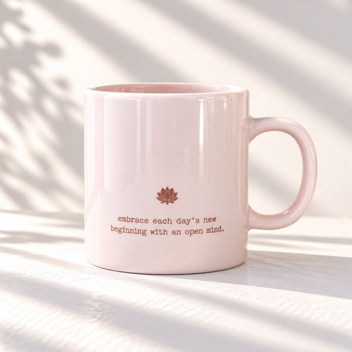 Glossy blush pink mug with a small lotus flower motif and the words “embrace each day’s new beginning with an open mind.”