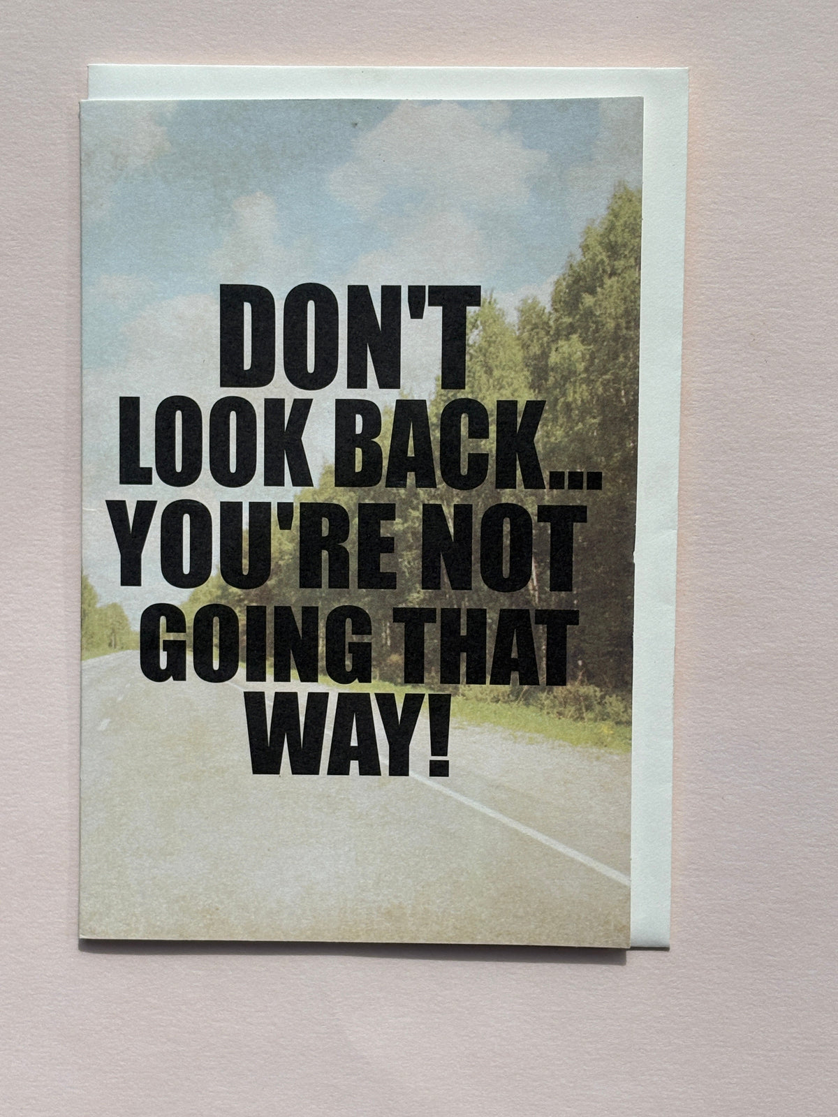 Greeting card with bold black text “Don’t look back, you’re not going that way” on a scenic road background