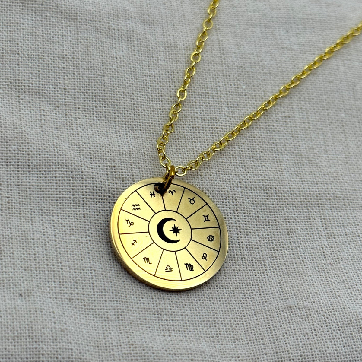 Gold astrology wheel necklace with zodiac symbols and crescent moon star pendant on a fine chain, styled on neutral fabric.