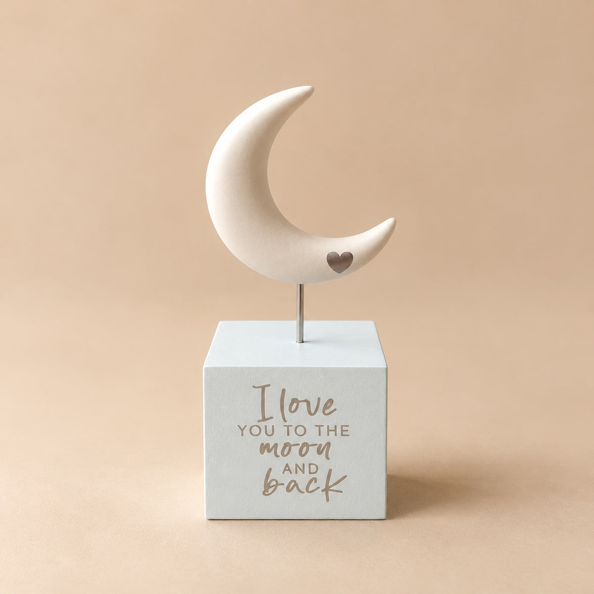 Cream crescent moon ornament with a small heart on a slim metal coloured stem above a pale square base printed “I love you to the moon and back”.
