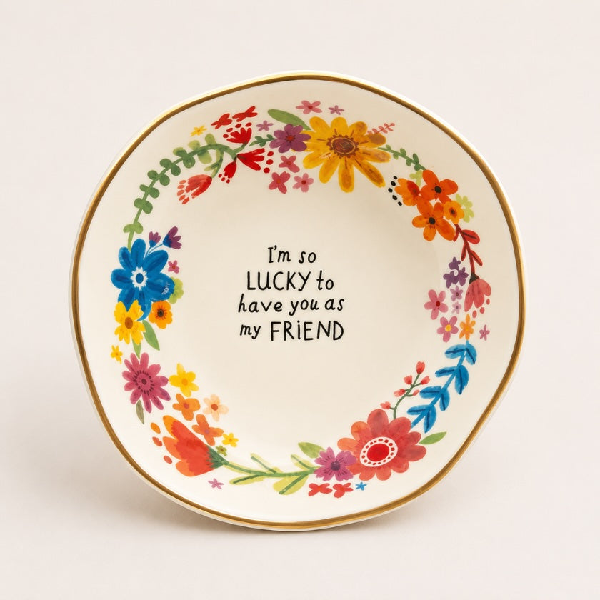 White ceramic trinket bowl with colourful floral wreath, gold rim detail and “I’m so lucky to have you as my friend” quote in the centre.