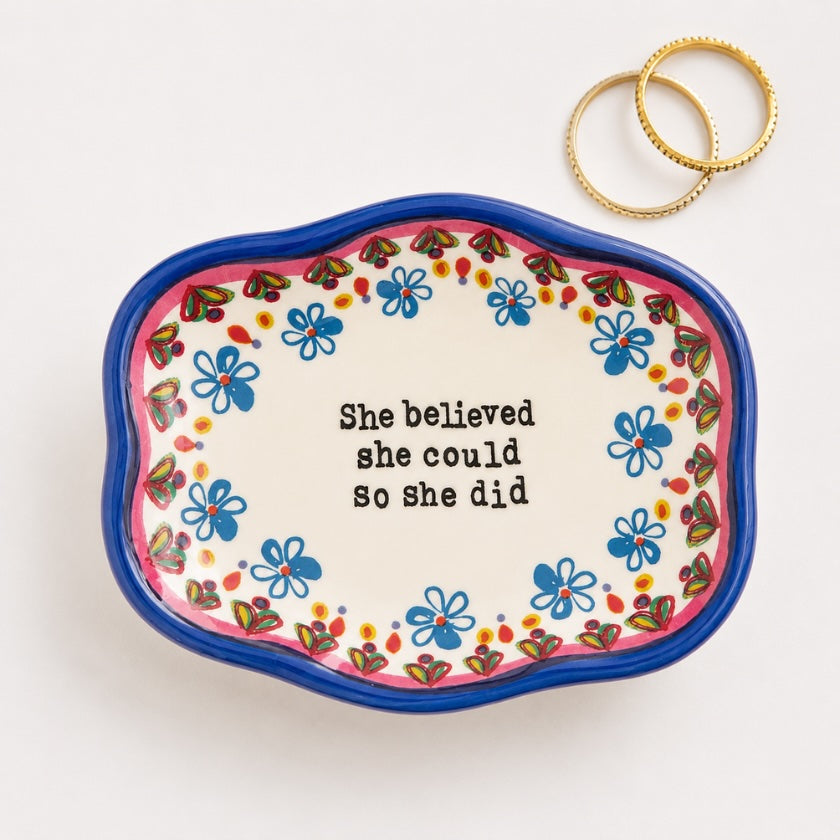 Hand painted style ceramic trinket bowl with blue floral border, inspirational “She believed she could so she did” quote and gold rings styled on a neutral background.