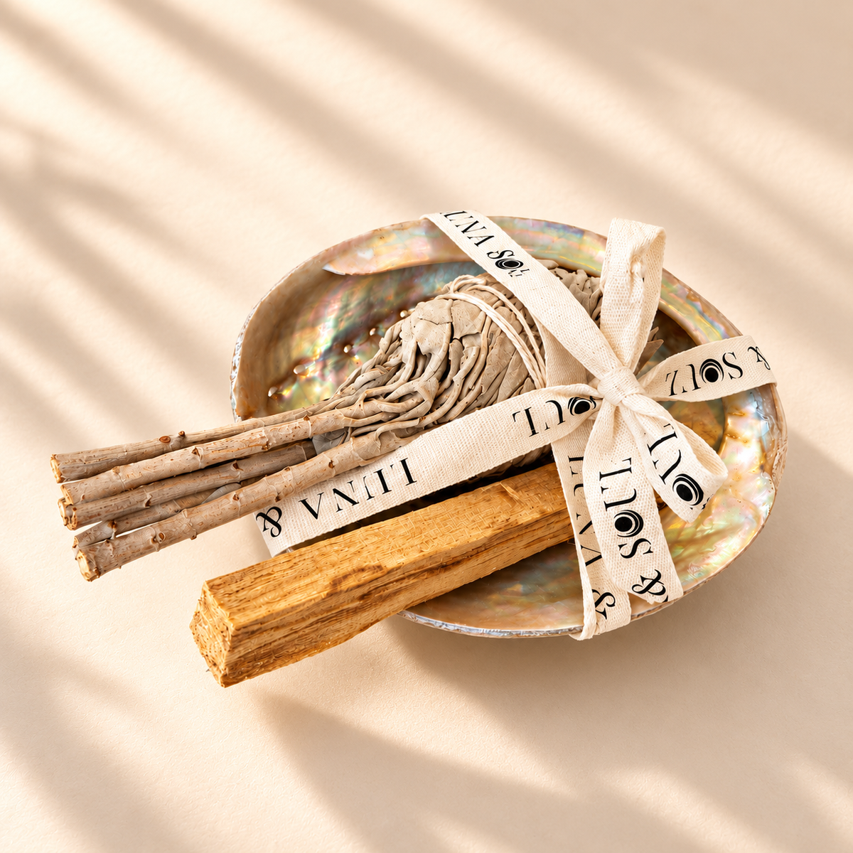 Luna &amp; Soul smudging gift set featuring an iridescent abalone shell, dried white sage bundle and natural Palo Santo wood stick, tied with branded cotton ribbon on a warm cream background.