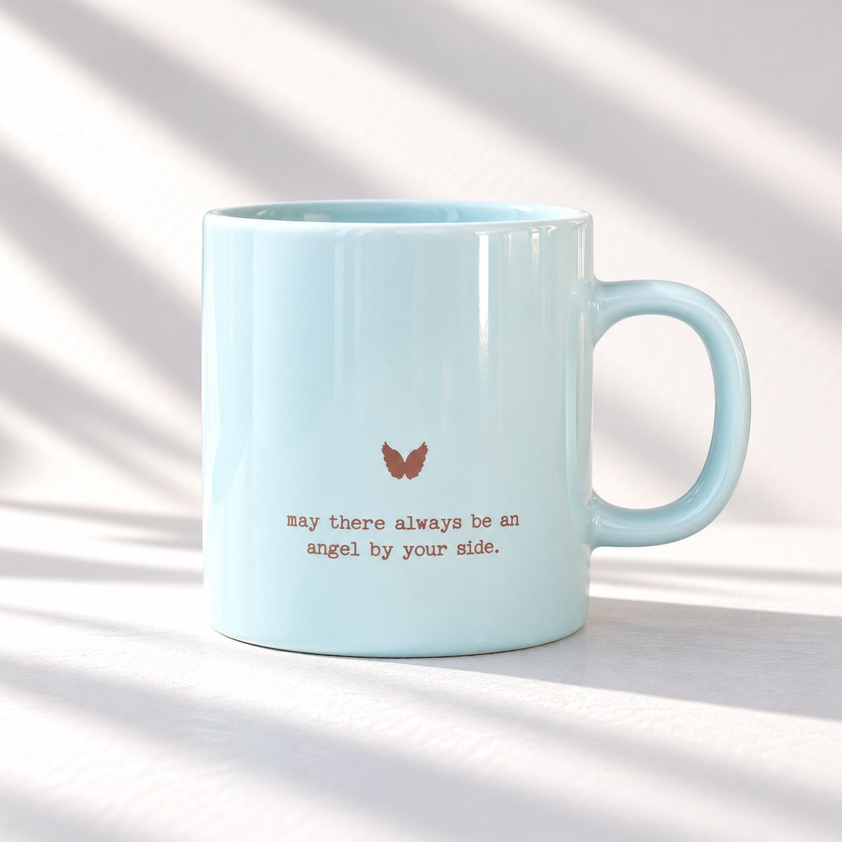 Pale blue glossy mug with a small metallic looking angel wings motif above “may there always be an angel by your side.”