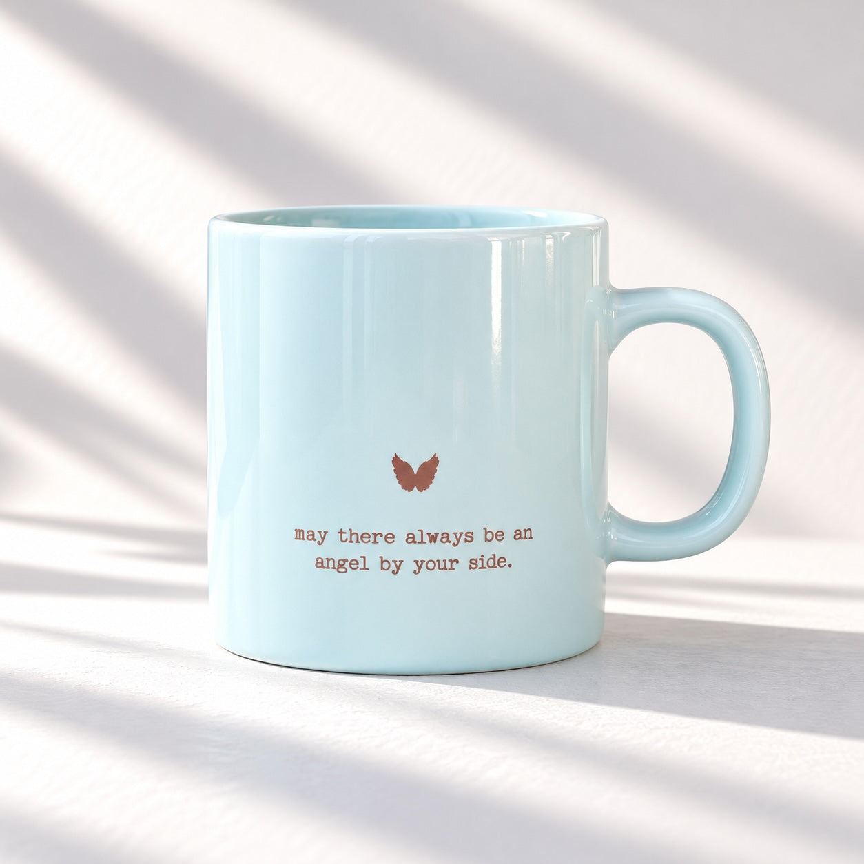Pale blue glossy mug with a small metallic looking angel wings motif above “may there always be an angel by your side.”