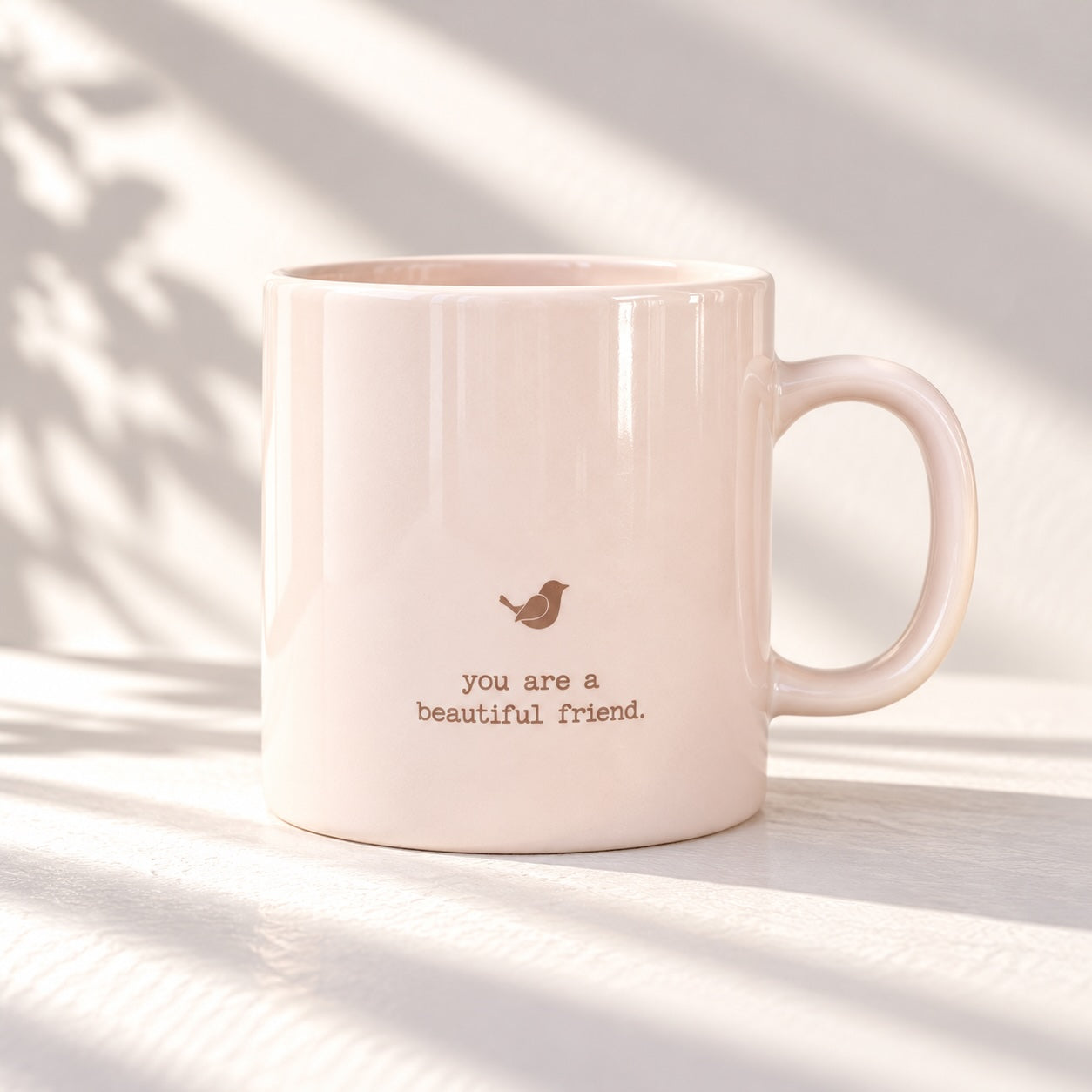 Glossy pale pink mug with a small bird motif above the words “you are a beautiful friend.”, handle on the right against a white background.