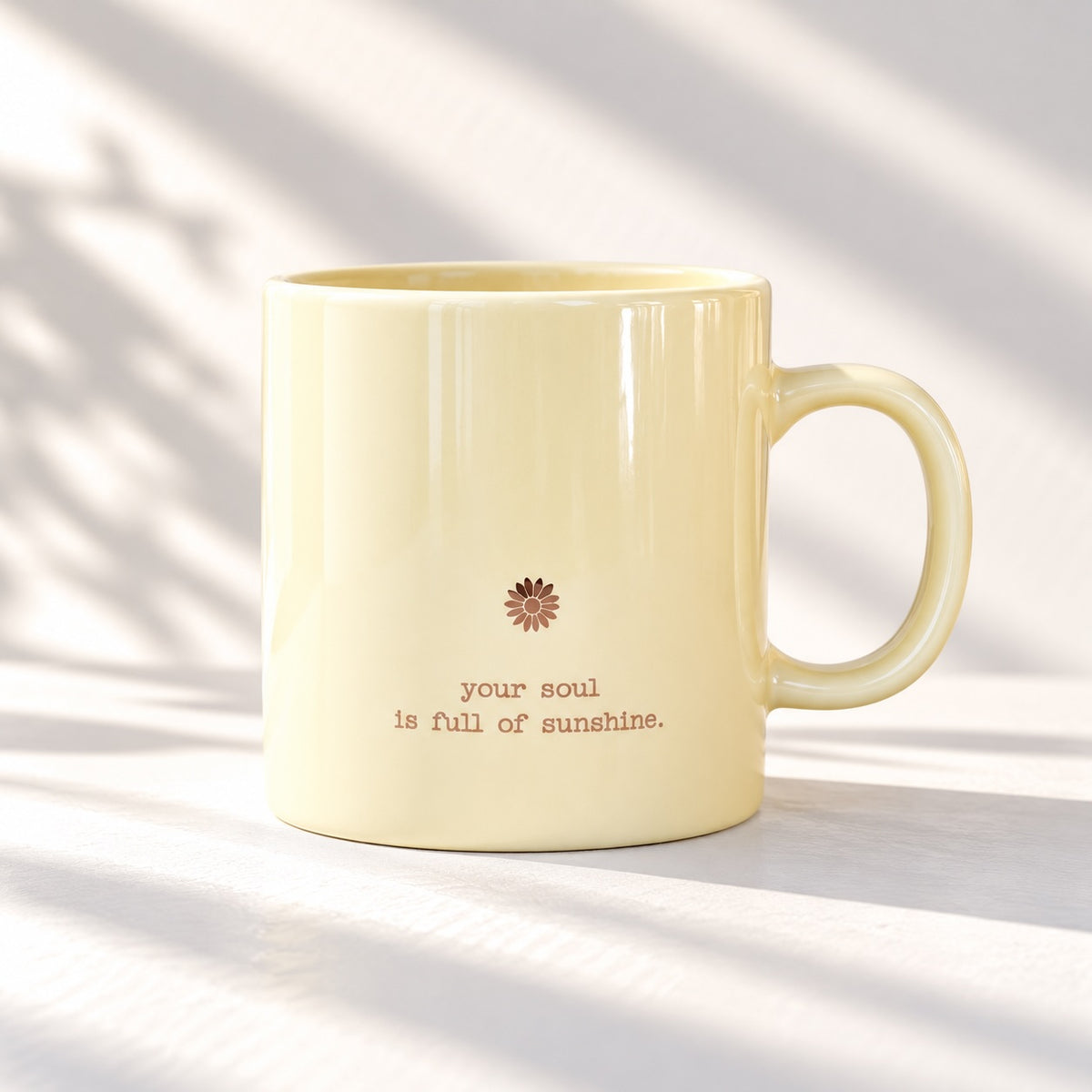 Glossy pale yellow mug with a small flower motif and “your soul is full of sunshine.” lettering, handle on the right, photographed in soft daylight.