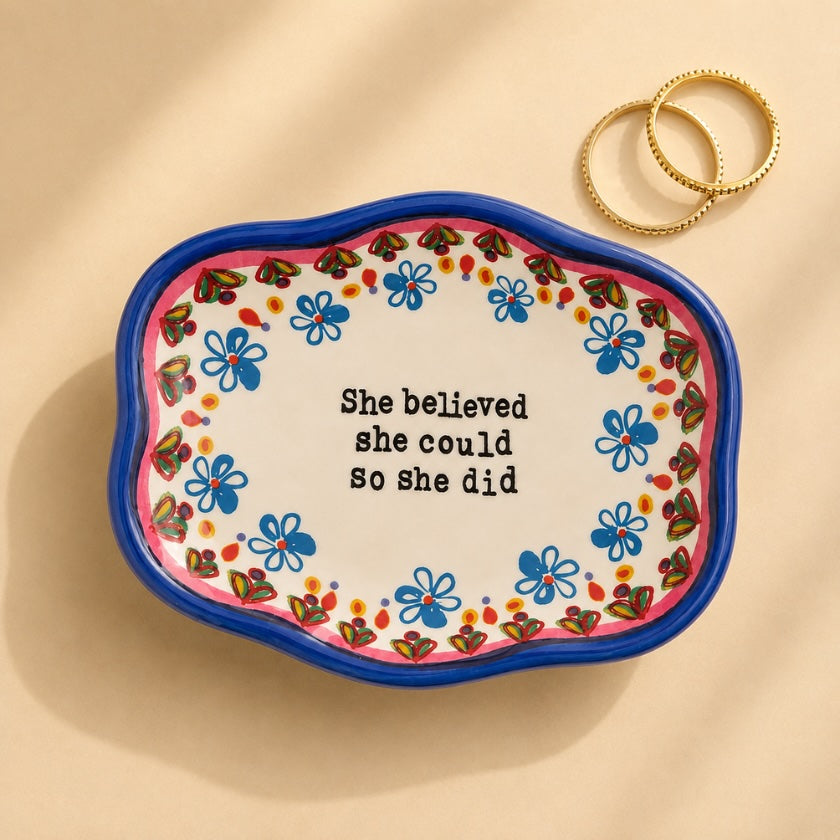 Hand painted style ceramic trinket bowl with blue floral border, inspirational “She believed she could so she did” quote and gold rings styled on a neutral background.