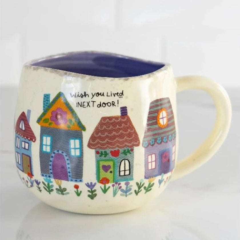 Cream artisan ceramic mug with colourful illustrated houses and flowers, a dark interior and the words “Wish you Lived NEXT door!”